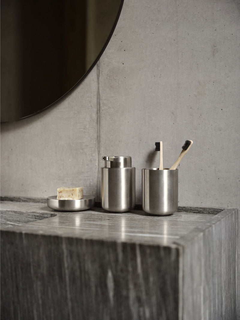 Three brushed stainless steel bathroom accessories arranged on a gray stone countertop against a concrete wall: a small round soap dish with a bar of soap, a cylindrical container, and a toothbrush holder with two toothbrushes.