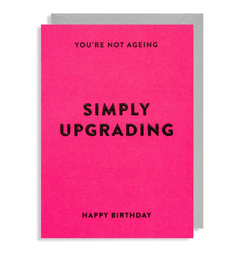 A pink birthday card with the text 'SIMPLY UPGRAADING HAPPY BIRTHDAY' in white capital letters, displayed on a table.