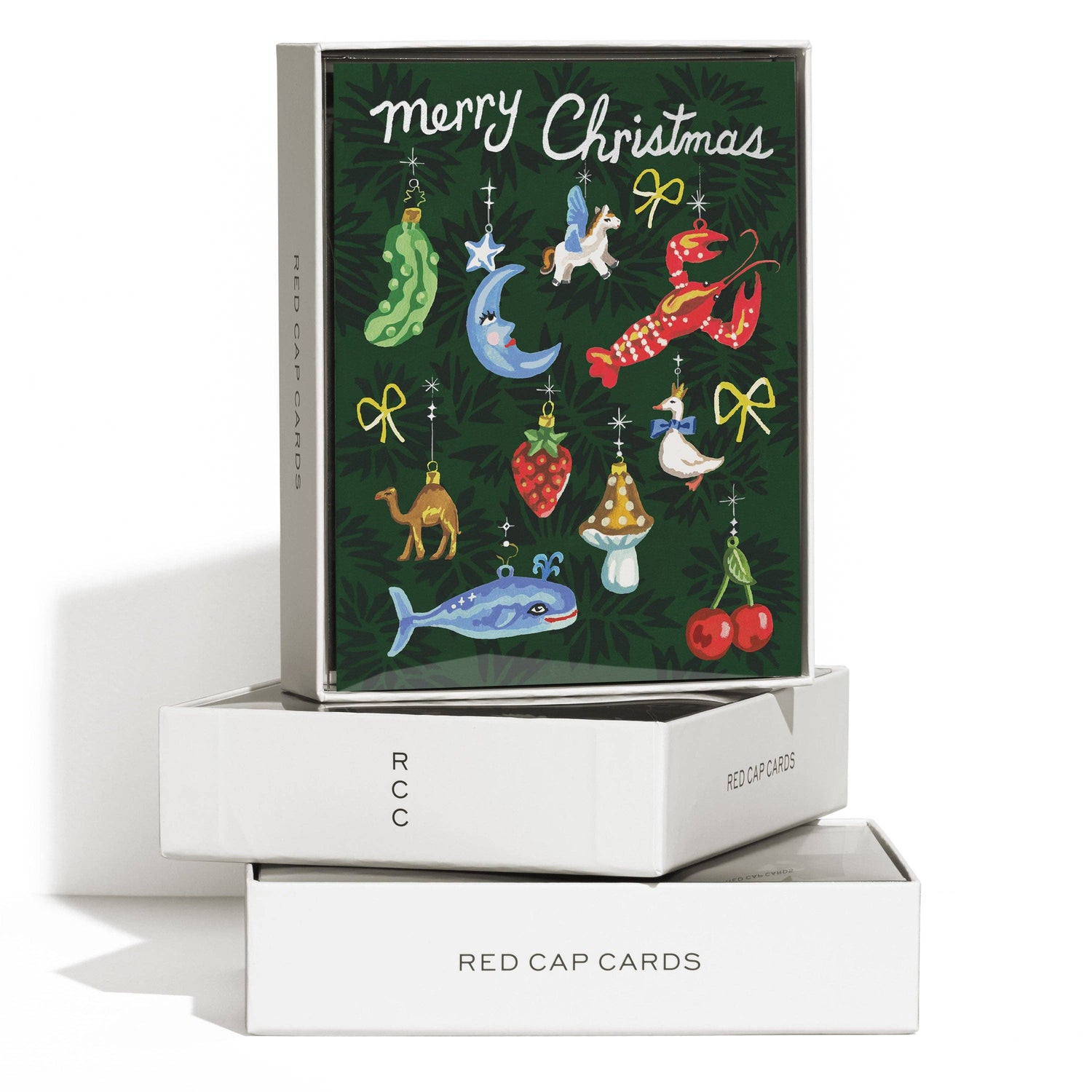 A Christmas greeting card set displayed on stacked white boxes. The top box shows a dark green card with colorful illustrated holiday ornaments and the words “merry Christmas.