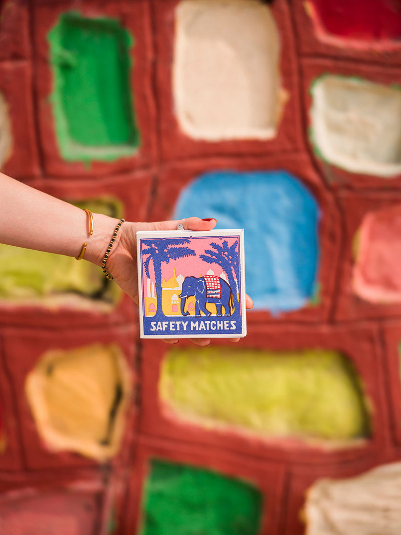 Person holding a safety matches box with an elephant design against a colorful, textured background.