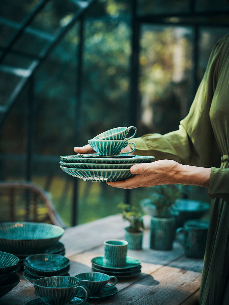 Person holding a stack of green ceramic tea cups and saucers in a blurred outdoor setting