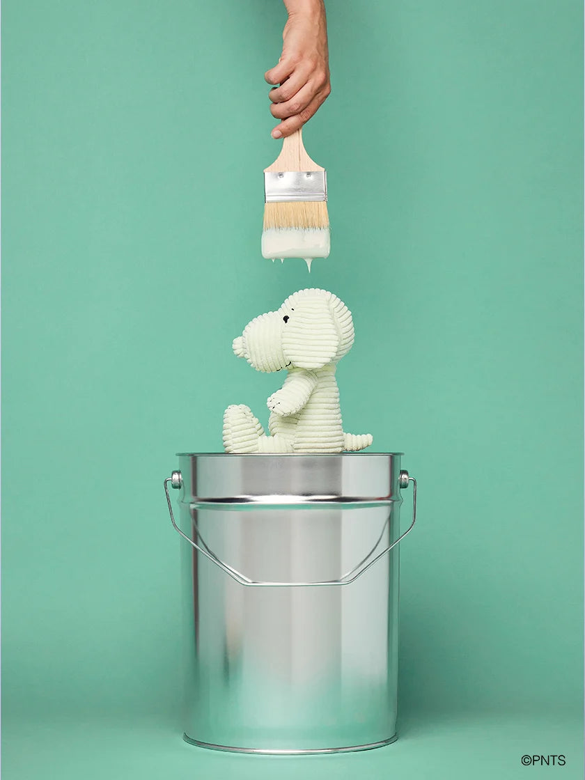 Toy snoopy dog being painted with a brush from a metal bucket against a green background.