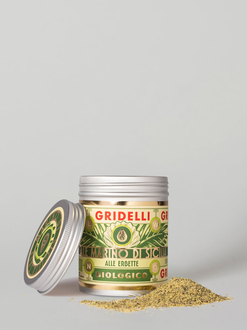 A jar of Gridelli Decoloro di Sigeno alle erbette biological hair conditioner with its silver lid leaning against it, with a small pile of the beige-green herbal powder spilled in front, on a light gray background.