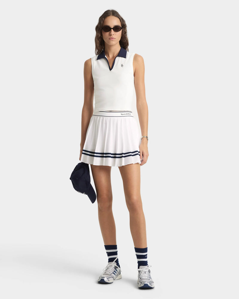 Woman wearing a white tennis skirt and top with navy accents on a white background