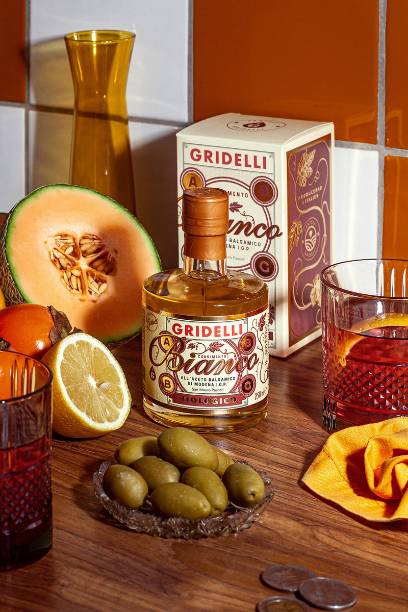 Gridelli Bianco bottle with a cantaloupe, lemon, olives, and coins on a wooden surface.