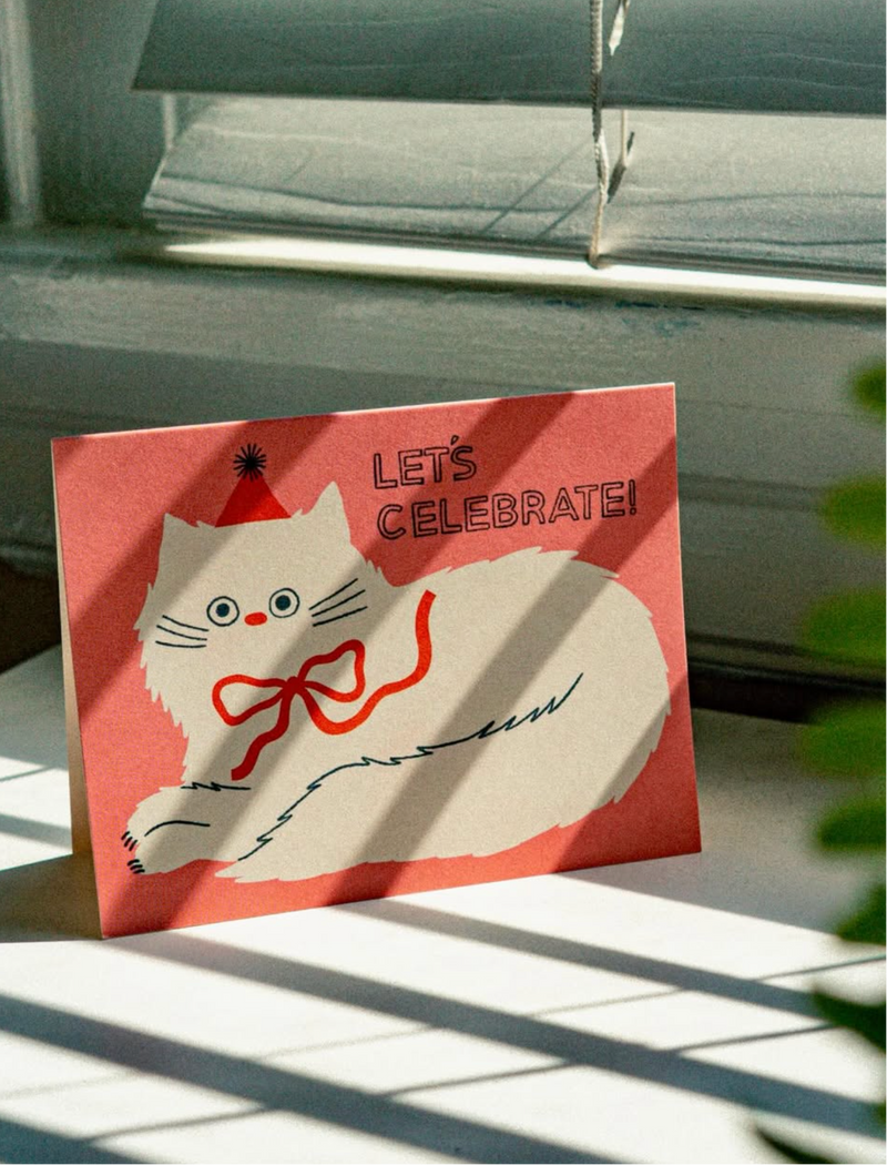 Card with a cat illustration and 'Let's Celebrate!' text on a surface with sunlight casting shadows.