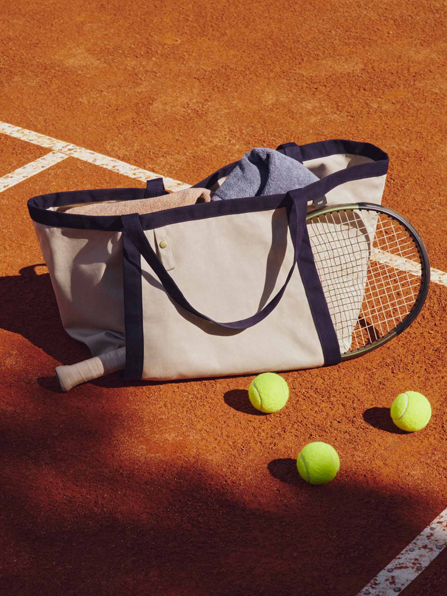 A canvas tennis tote bag with navy blue trim sits on a clay tennis court, with a tennis racket tucked inside, a towel draped over the top, and three yellow tennis balls nearby.