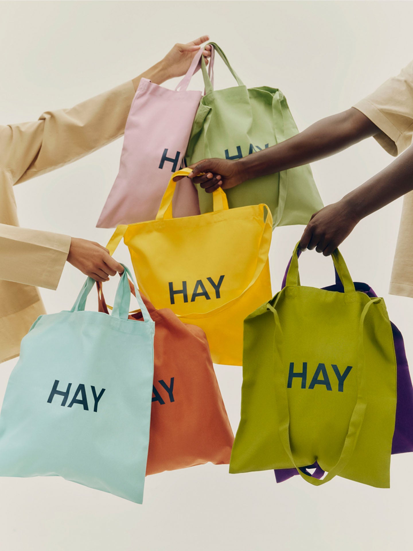 Six reusable cotton tote bags in pastel and bright colors, each printed with the word 'HAY', held by multiple hands against a clean off-white background.