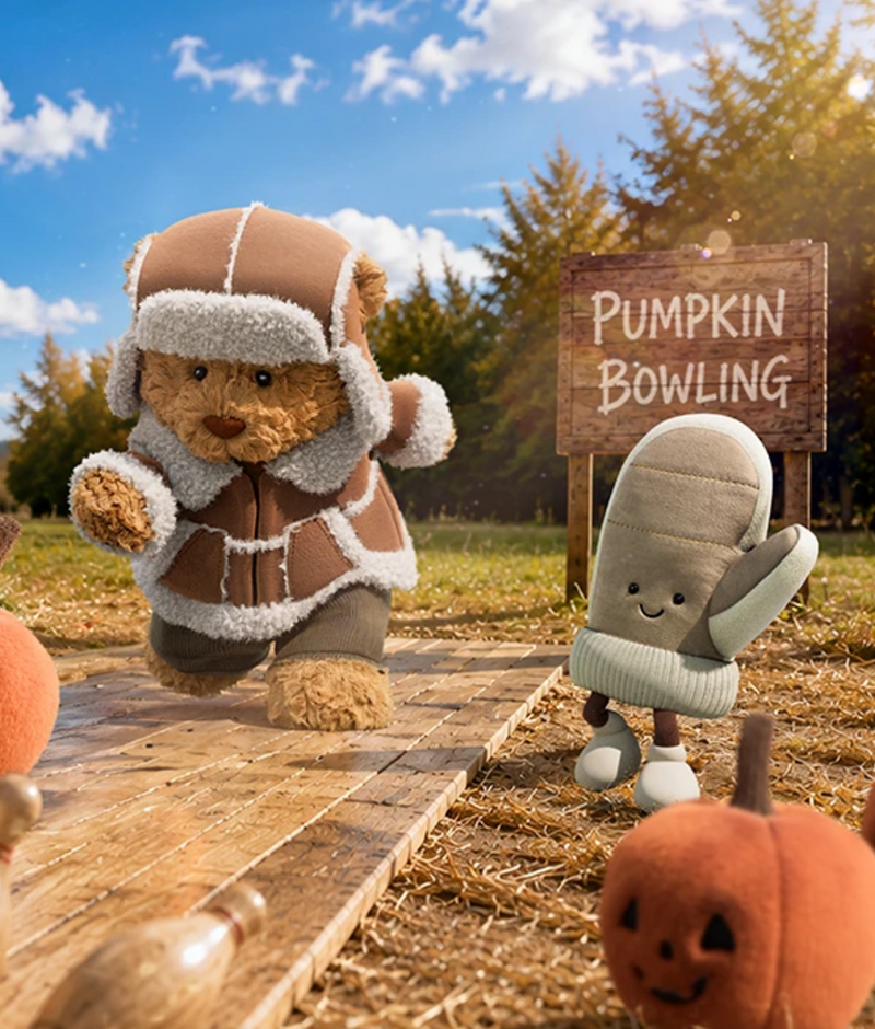 A teddy bear wearing a brown and beige fuzzy aviator-style jacket and hat stands on a wooden path outdoors, next to a gray mitten character. A wooden sign reads “PUMPKIN BOWLING,” with pumpkins and trees in the background under a blue sky.