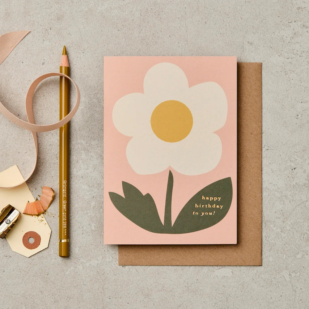 Birthday greeting card with a flower design and 'happy birthday to you!' text, placed on a textured surface with stationery items.