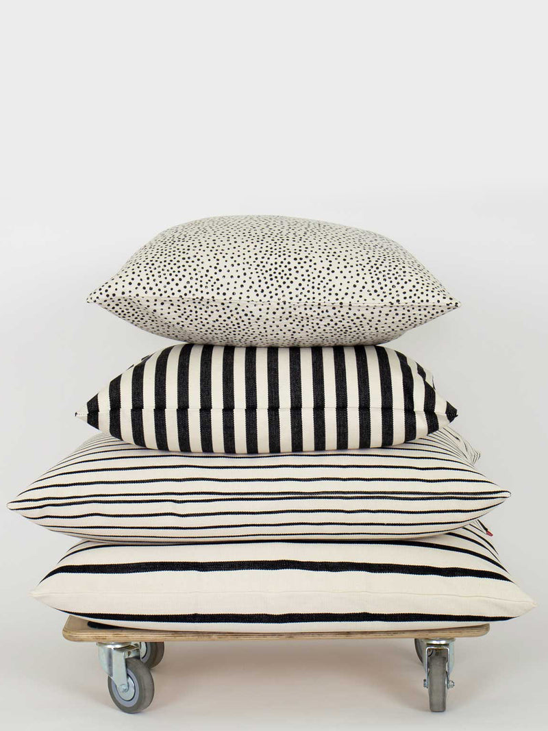 A square cushion made from off-white cotton with black hand-printed flecks or irregular little dots on top of a a same sized black and white striped square cushion and two bigger black and white striped cushions suitable for home decoration such as couches, settees, sofas and armchairs, stacked in a pile on a low table, photographed against a grey studio background