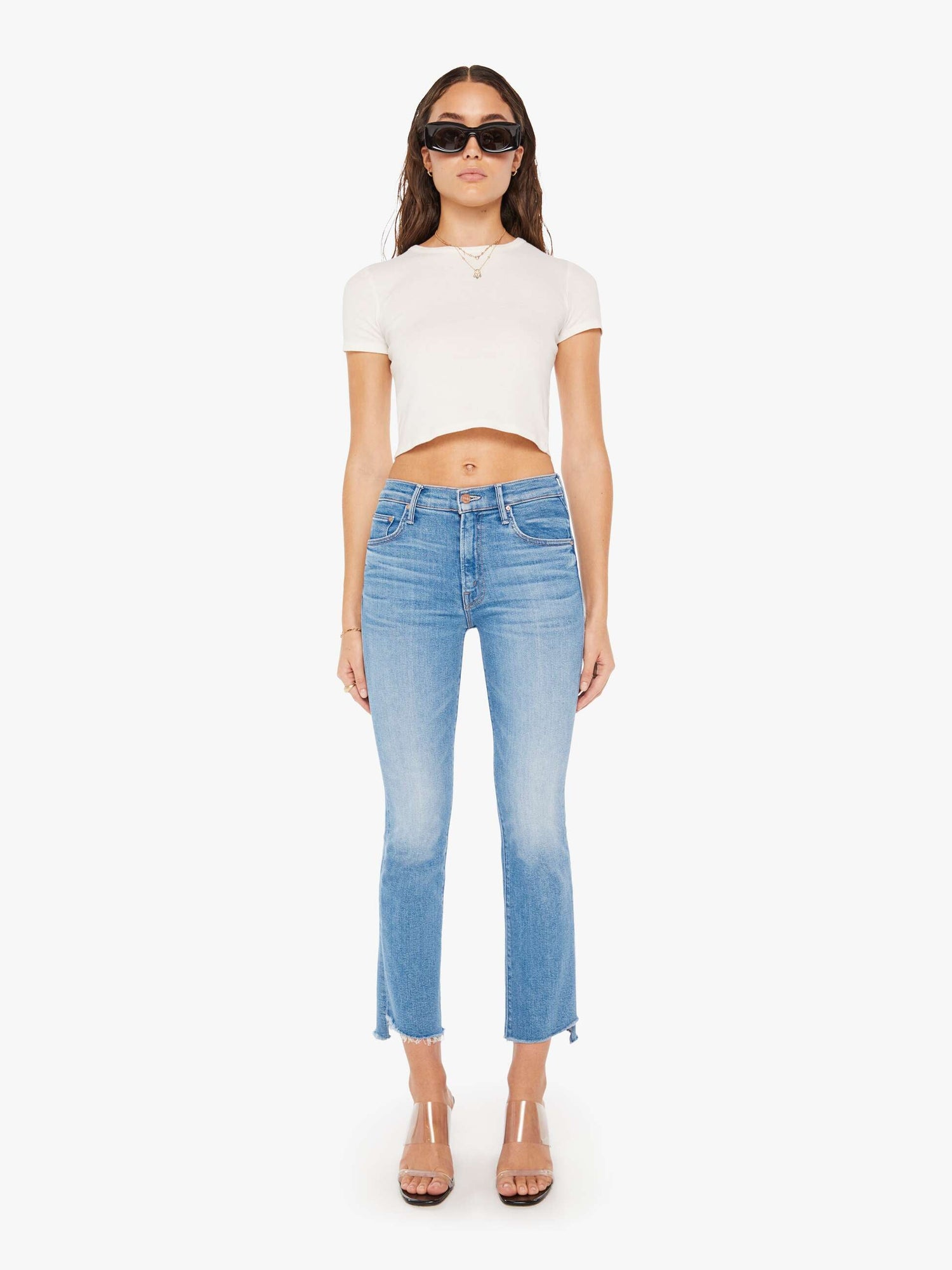 A female model wearing high-waisted blue bootcut jeans with a frayed step-hem and a mid-blue wash, paired with a white crop top.