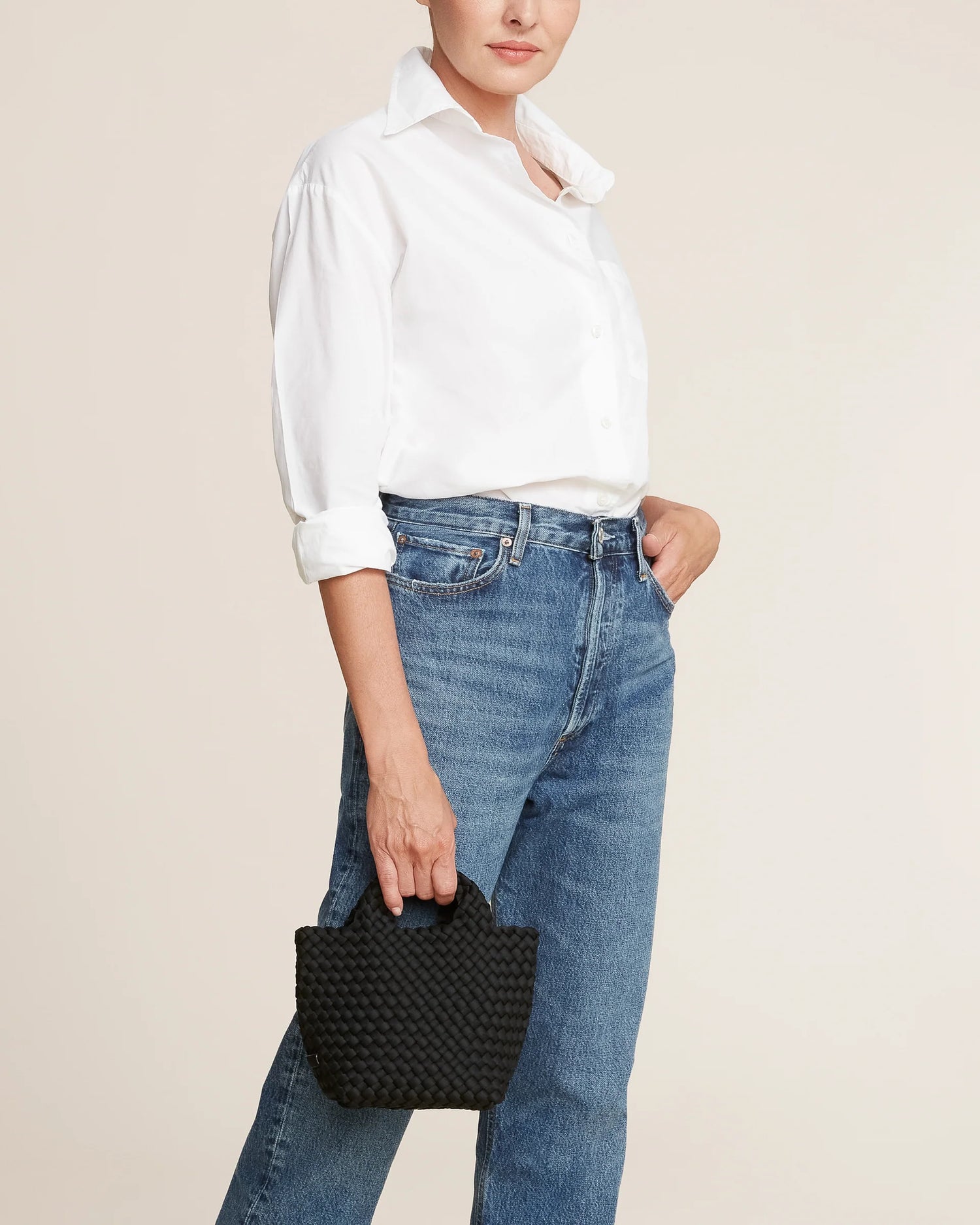 Woman wearing a white shirt and blue jeans, holding a black handbag by Naghedi against a beige background