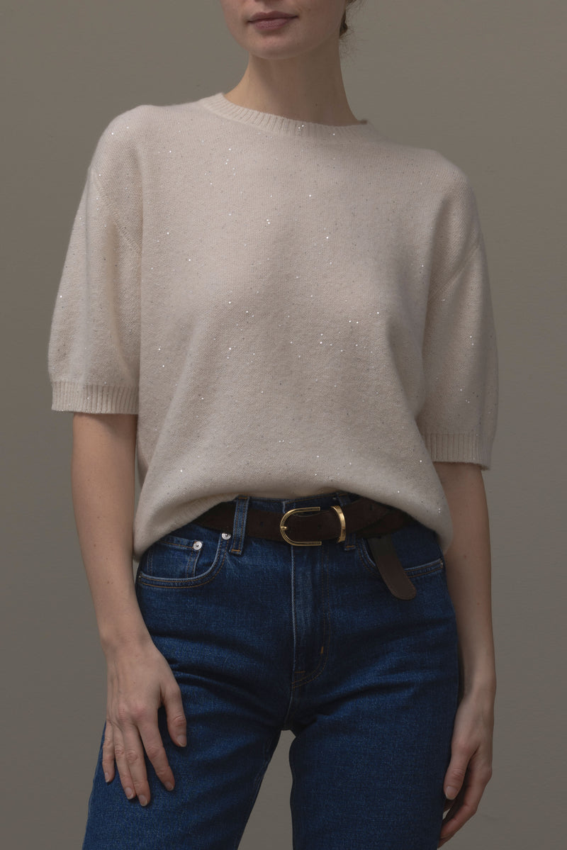 A model wearing a short-sleeve cream knit sweater with subtle shimmering speckles, tucked little into dark blue jeans with a brown leather belt.