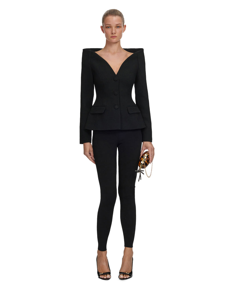 A model stands against a plain white background wearing a black tailored outfit consisting of a structured long-sleeve blazer with a deep V-neck and matching slim-fit pants. She is holding a small patterned clutch and wearing black strappy heels.