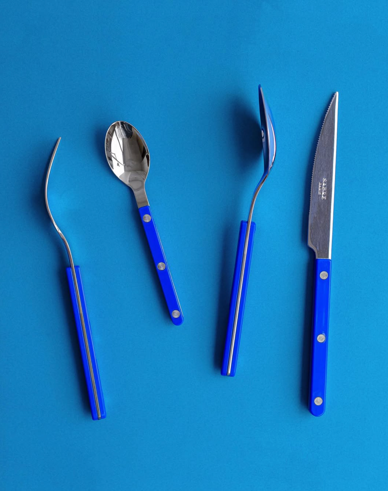 Set of blue-handled cutlery including a fork, tea spoon, soup spoon and knife on a blue background.