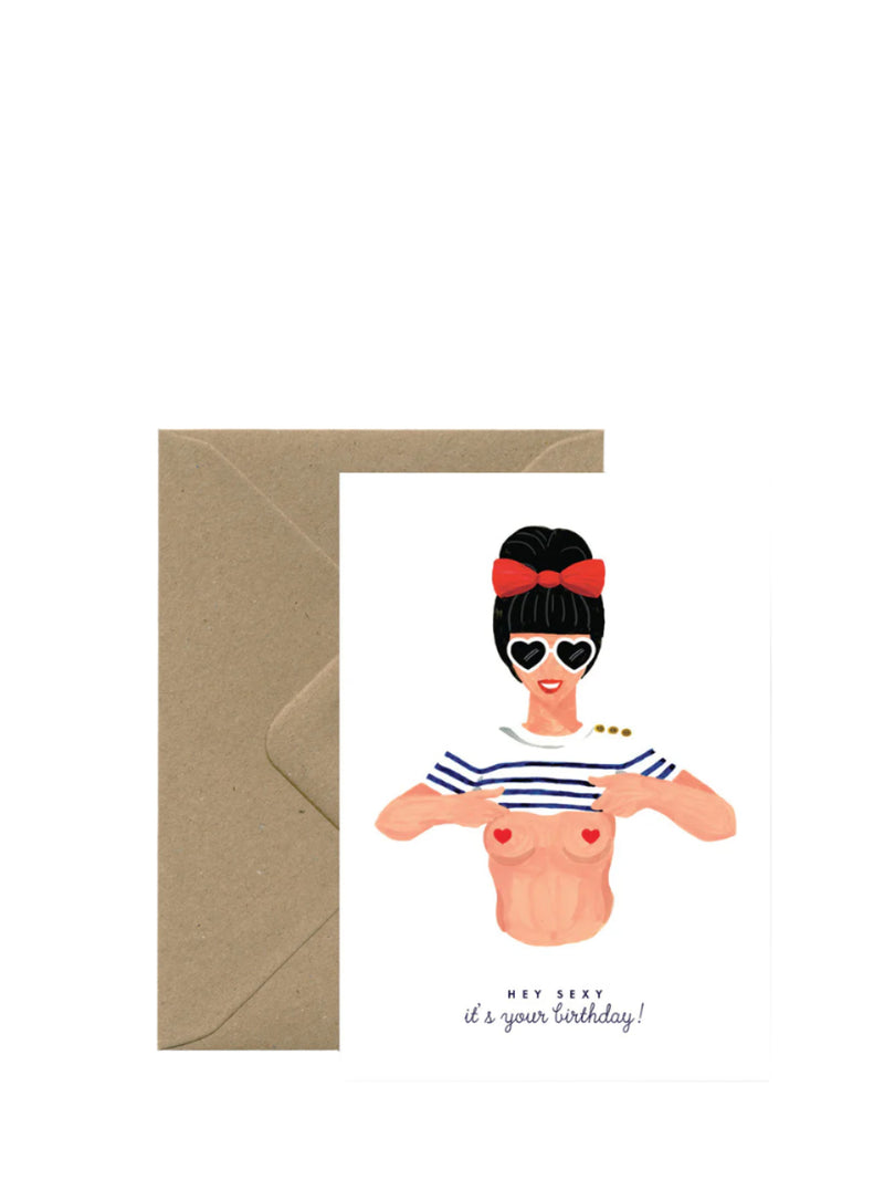A birthday card with an illustration of a woman wearing sunglasses and a striped top, with a text that reads 'Happy Birthday! Now you're old!' The card is accompanied by a brown envelope.