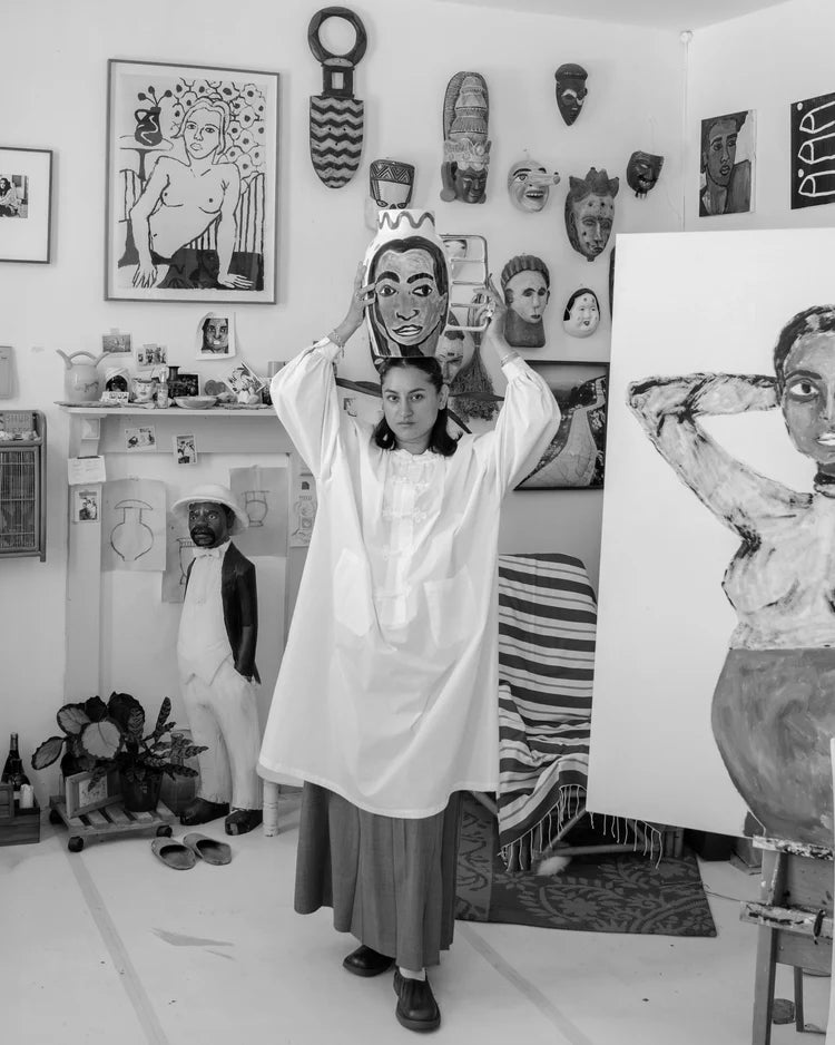 The artist in a white dress holding a vase with her design in an art studio with various artworks and sculptures.