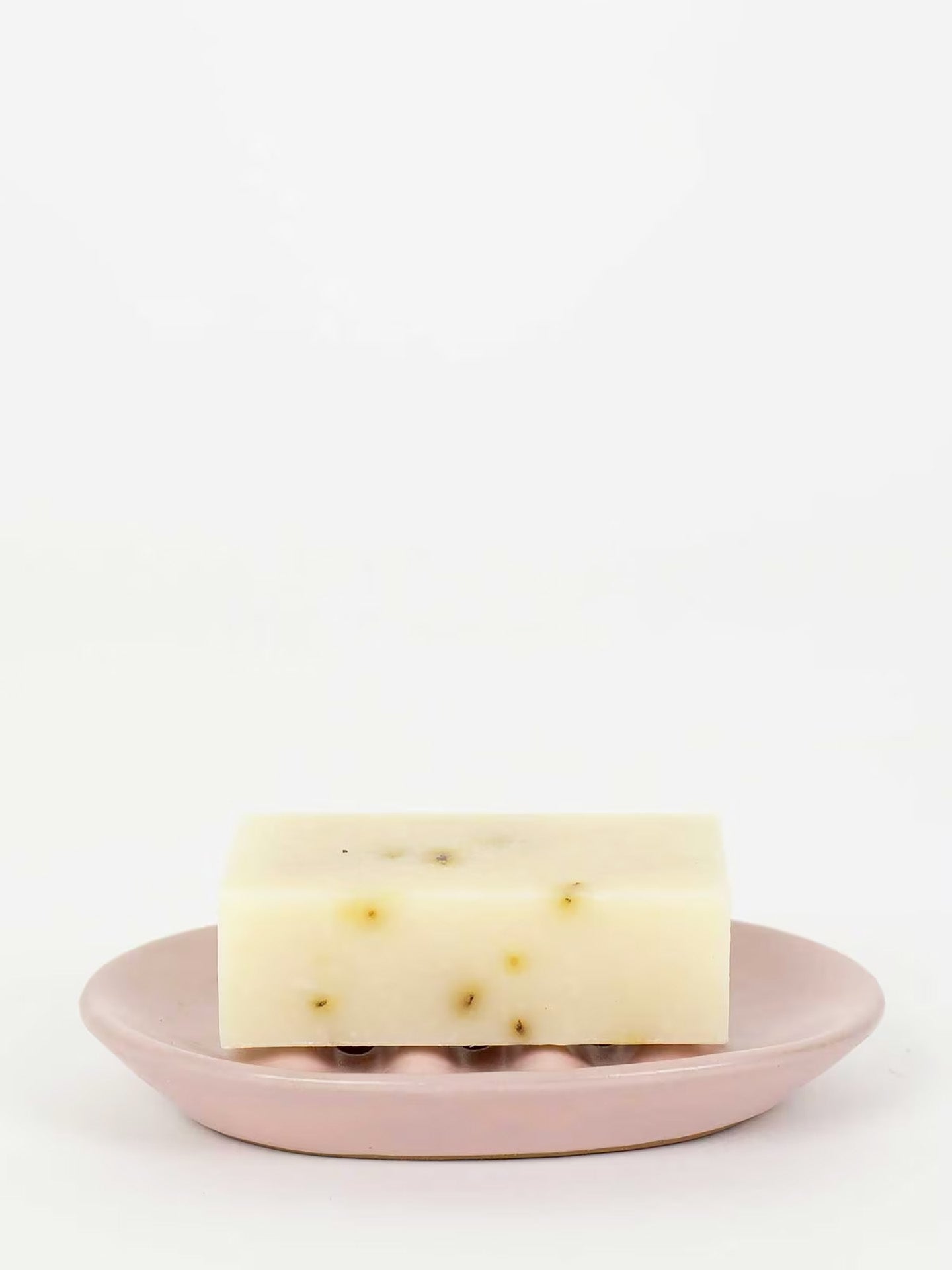 A rectangular bar of pale cream soap with small dark specks, resting on a dusty pink ceramic soap dish against a plain white background.