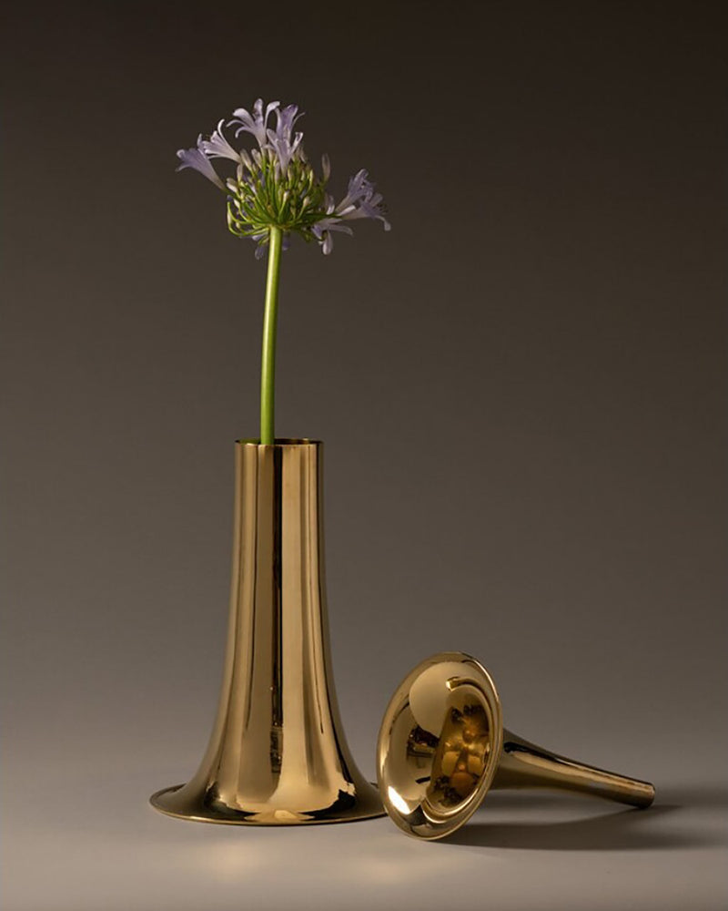 A polished gold-coloured metal horn-shaped vase holding a single purple flower, with similar, but smaller and slimmer golden metal flower vase lying beside it on a dark gray studio background.