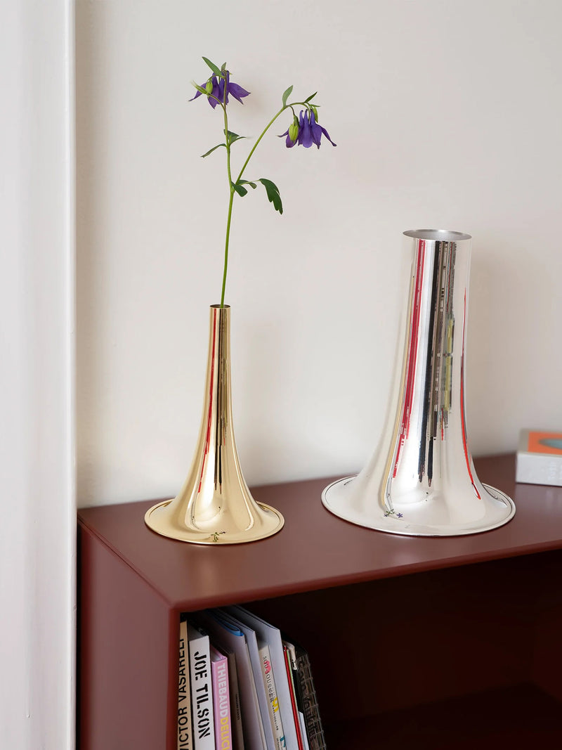 Two modern bell-shaped metal vases on a dark brown wooden shelf against a white wall, one in polished gold and one in polished silver, with purple flowers in the gold vase.