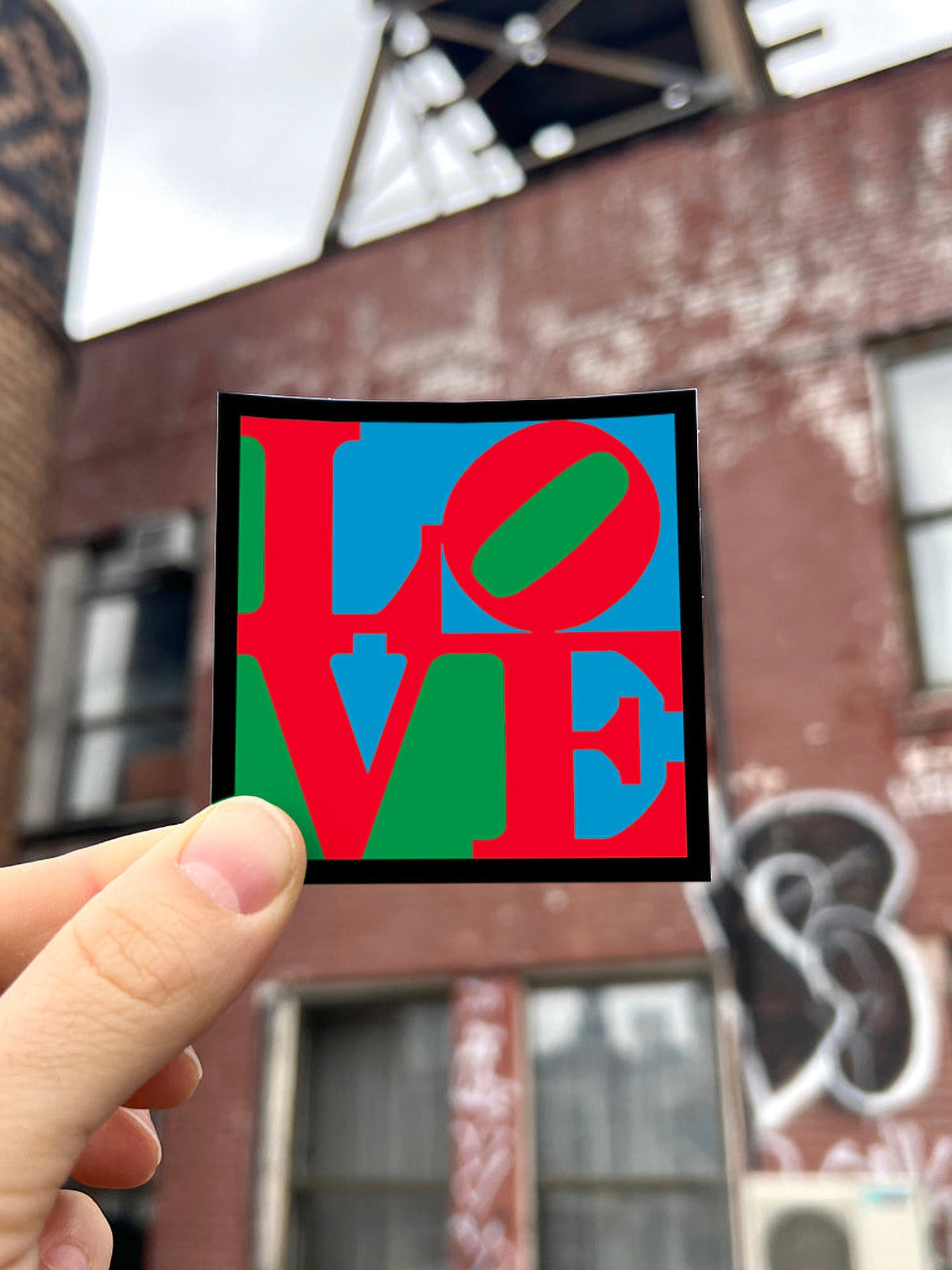 Colorful 'LOVE' graphic sticker held up against a blurred urban background