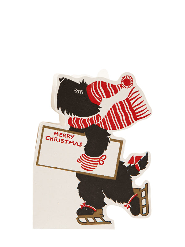 A vintage-style Christmas cutout featuring a black dog wearing a red and white striped knit hat and scarf while holding a sign that reads “MERRY CHRISTMAS.”.