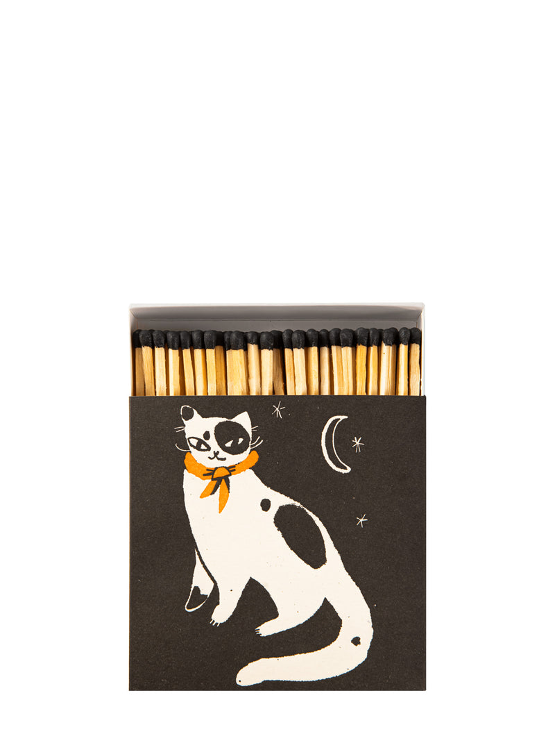 A matchbox with a dark brown cover featuring a white cat with black patches and an orange scarf, alongside a crescent moon and stars. The wooden matches with black tips are visible at the top.