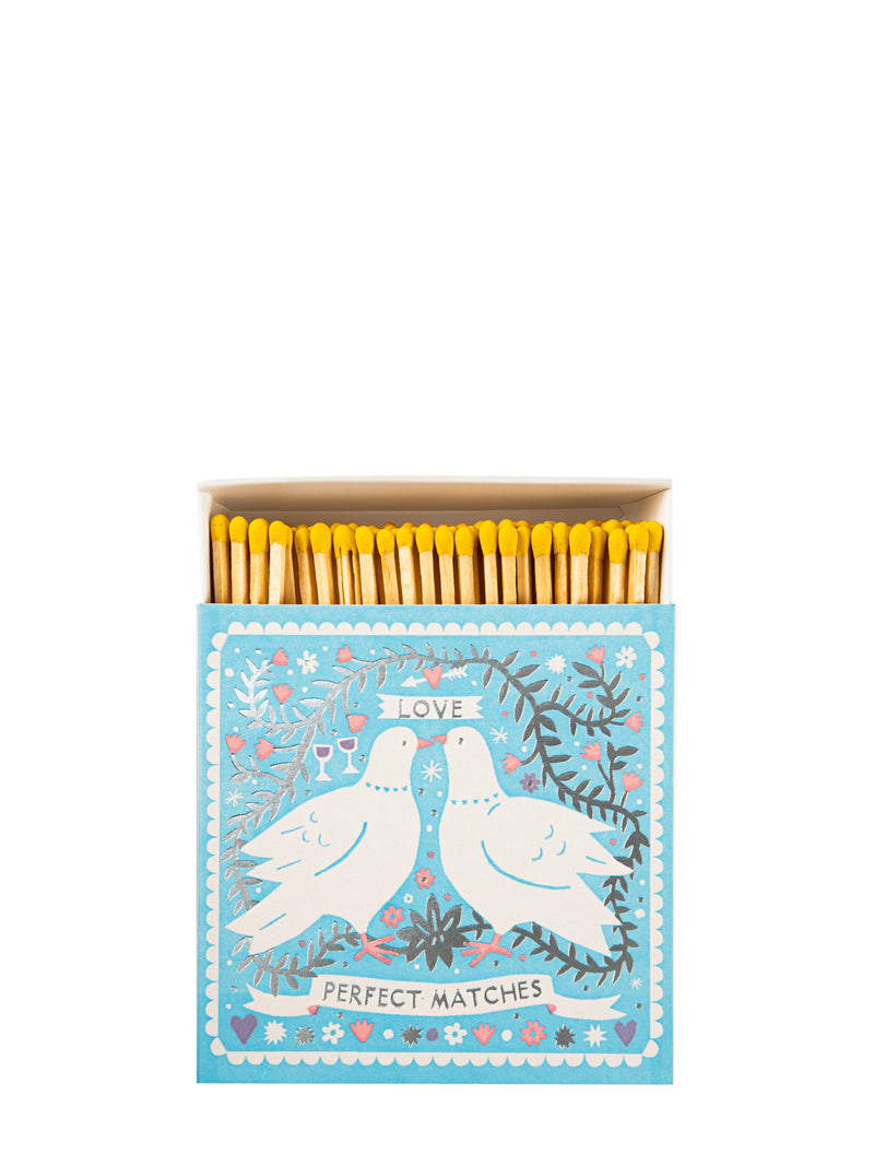 A decorative matchbox filled with yellow matches, shown against a plain white background. The matchbox lid features an illustration of two white doves facing each other, surrounded by floral and leafy details, with the words 'LOVE' and 'PERFECT MATCHES' on banners.