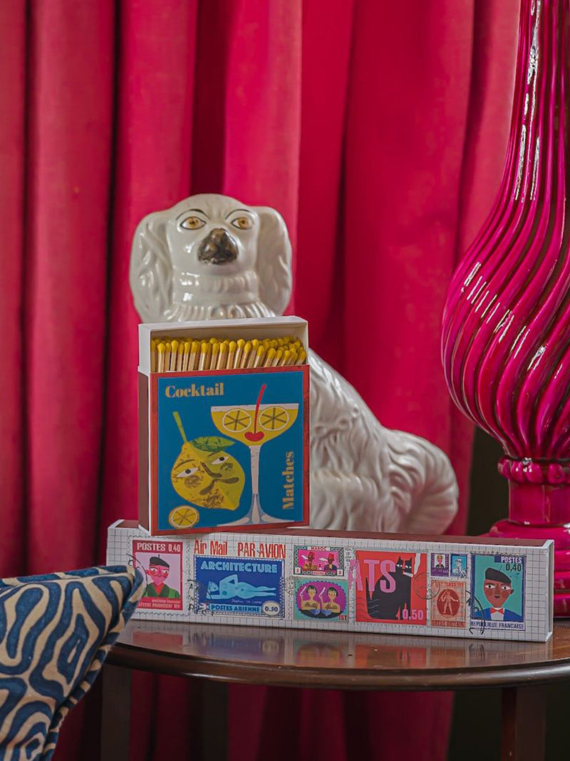 Decorative items including a ceramic dog, matchbox, and colorful stamps on a table with a pink curtain background.
