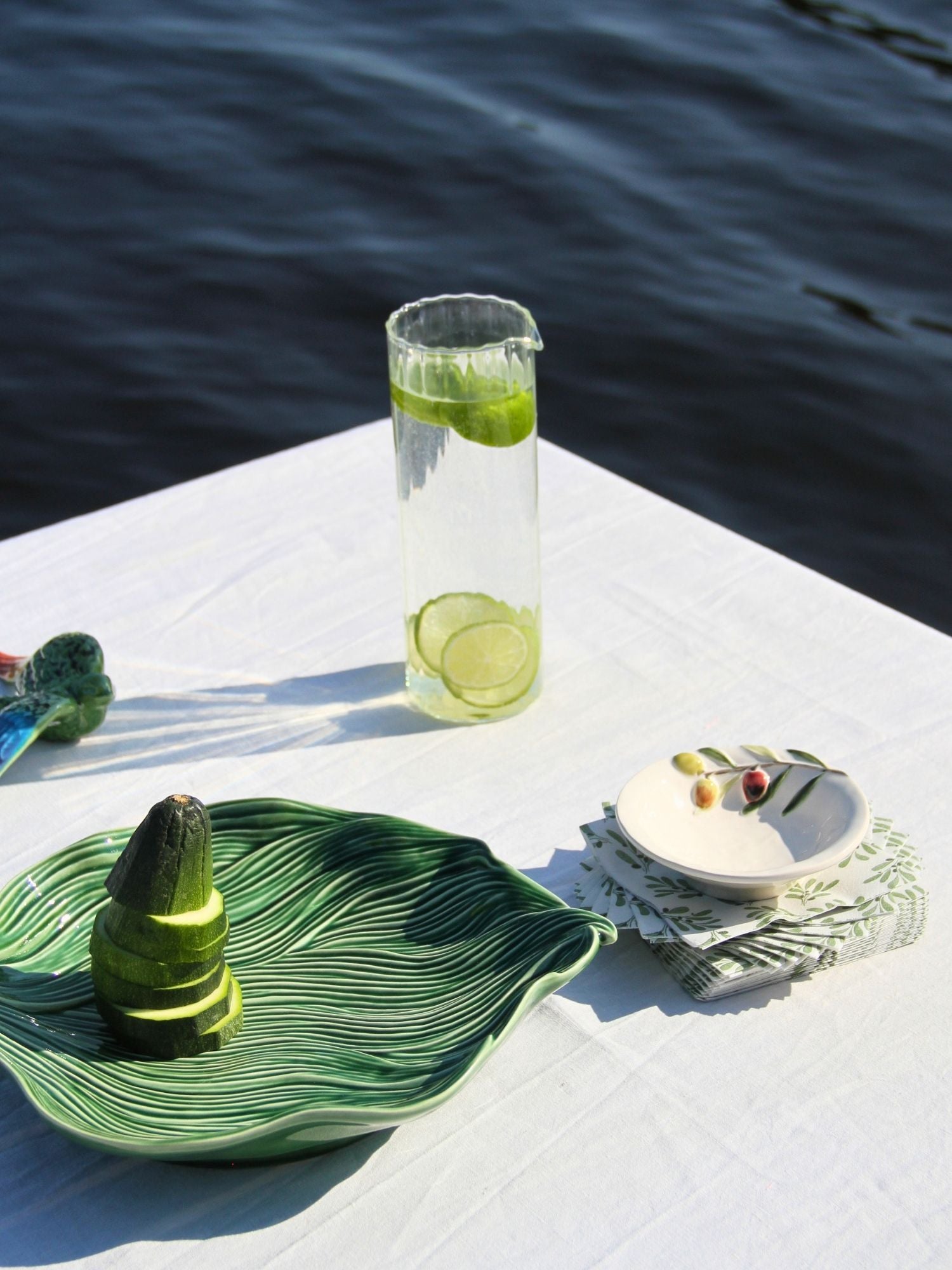 A styled outdoor table setting with a tall clear glass pitcher containing lime slices, a green leaf-shaped plate holding a cucumber, and a small white dish on a folded napkin, all set against dark blue water.