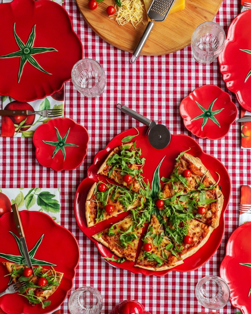 Red pizza pan with a pizza on a checkered tablecloth, surrounded by red tomato-shaped plates.