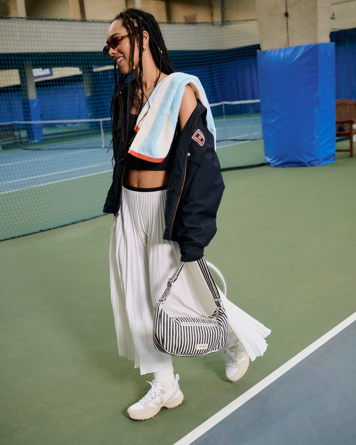 Person on a tennis court wearing a black jacket, white pleated skirt, and carrying a striped bag.