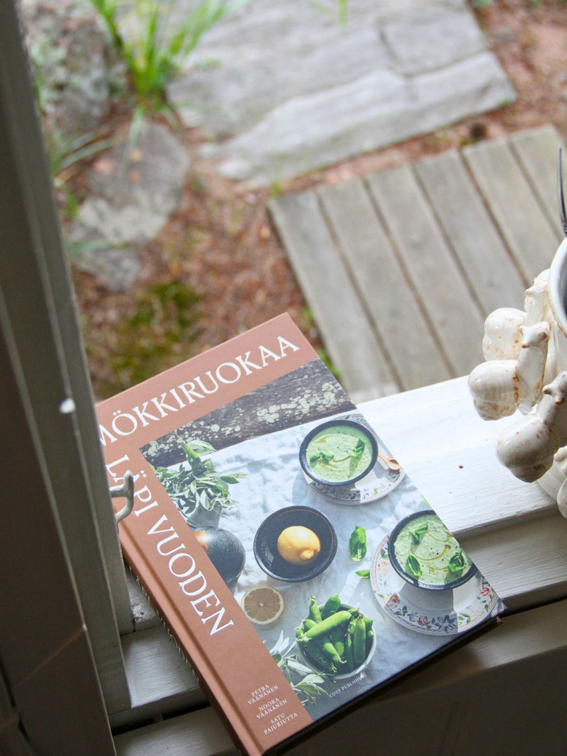 A cookbook titled 'MÖKKIRUOKAA LIPPUVUODEN' resting on a white windowsill with an outdoor garden background visible through the glass.