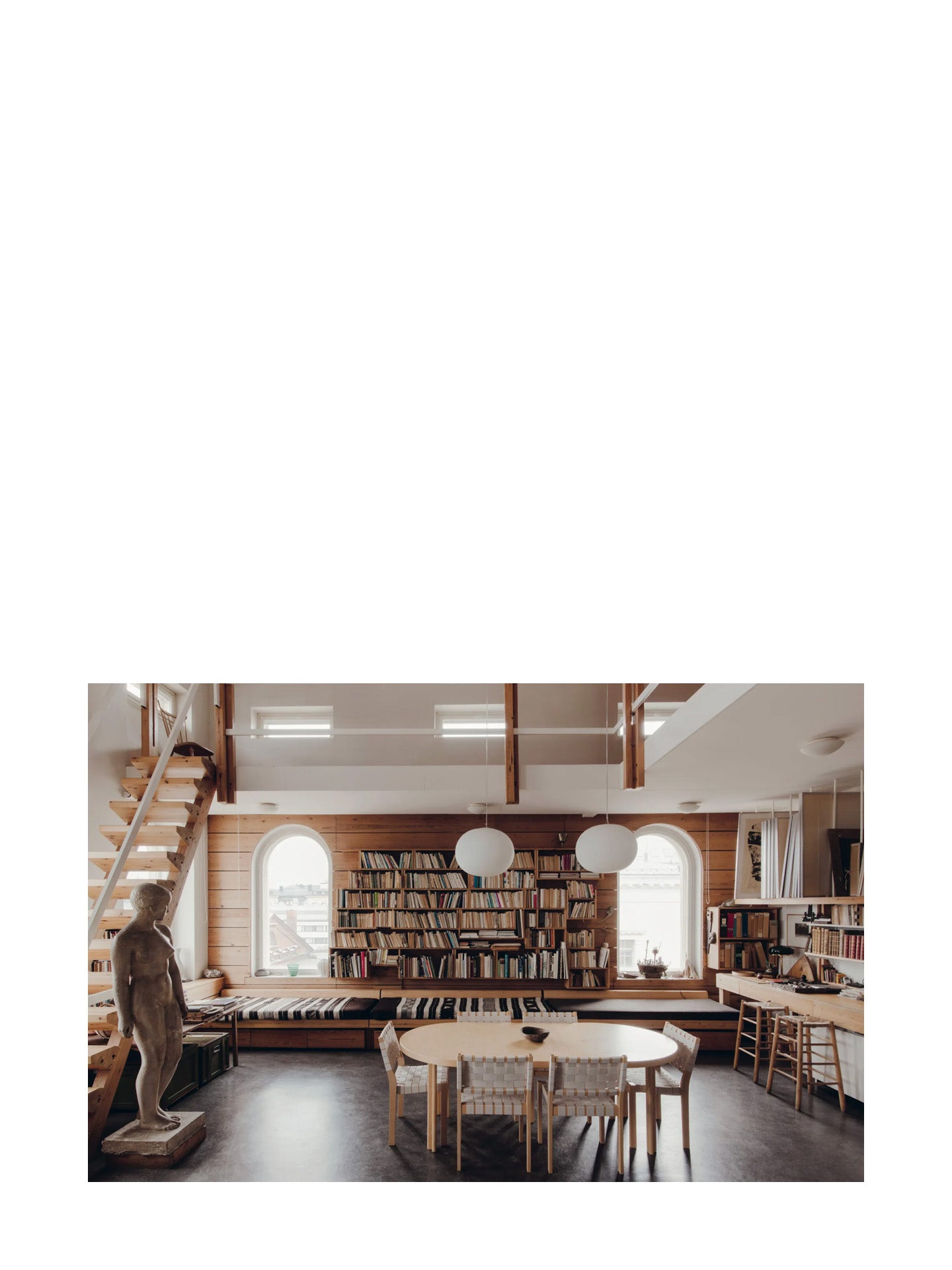 Atelier interior design of Tove Jansson's studio home with a arched windows, bookshelf, Artek dinner table, and chairs and stairs leading up the mezzanine next to a life-sized realistic sculpture of a woman standing.