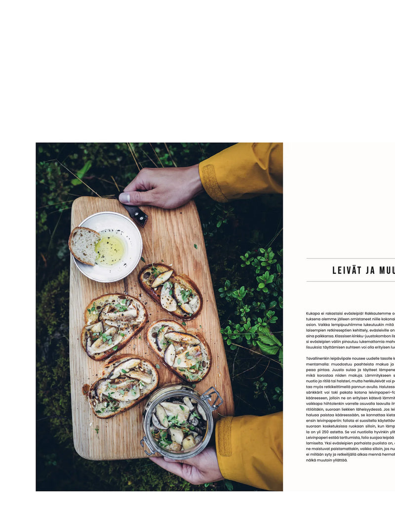 Person holding a wooden board with mushroom bruschettas and a small bowl of herbal oil outdoors of a lingonberry bush.