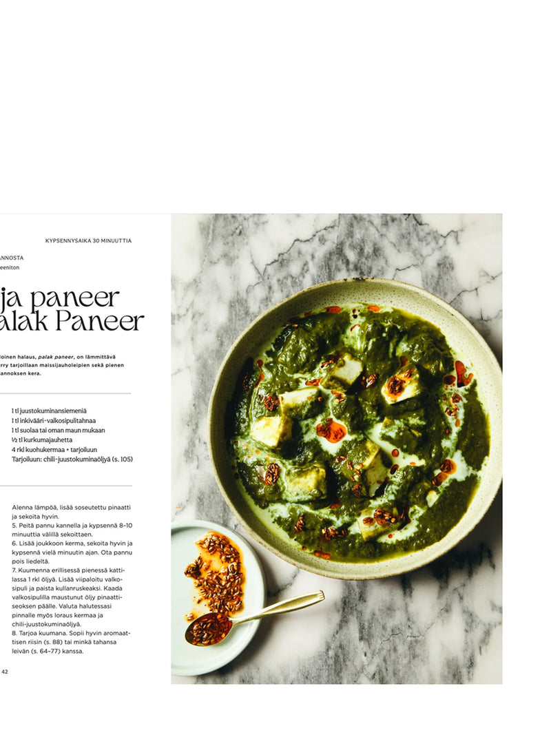 A partially shown book spread of a recipe for a green palak paneer dish with the recipe on the left page and a prepared food on a above photograped image featuring a chili oil speckled paneer in a round beige bowl on a marble surface, with a small dish of sesame seed chili oil and tea spoon on the lower left.
