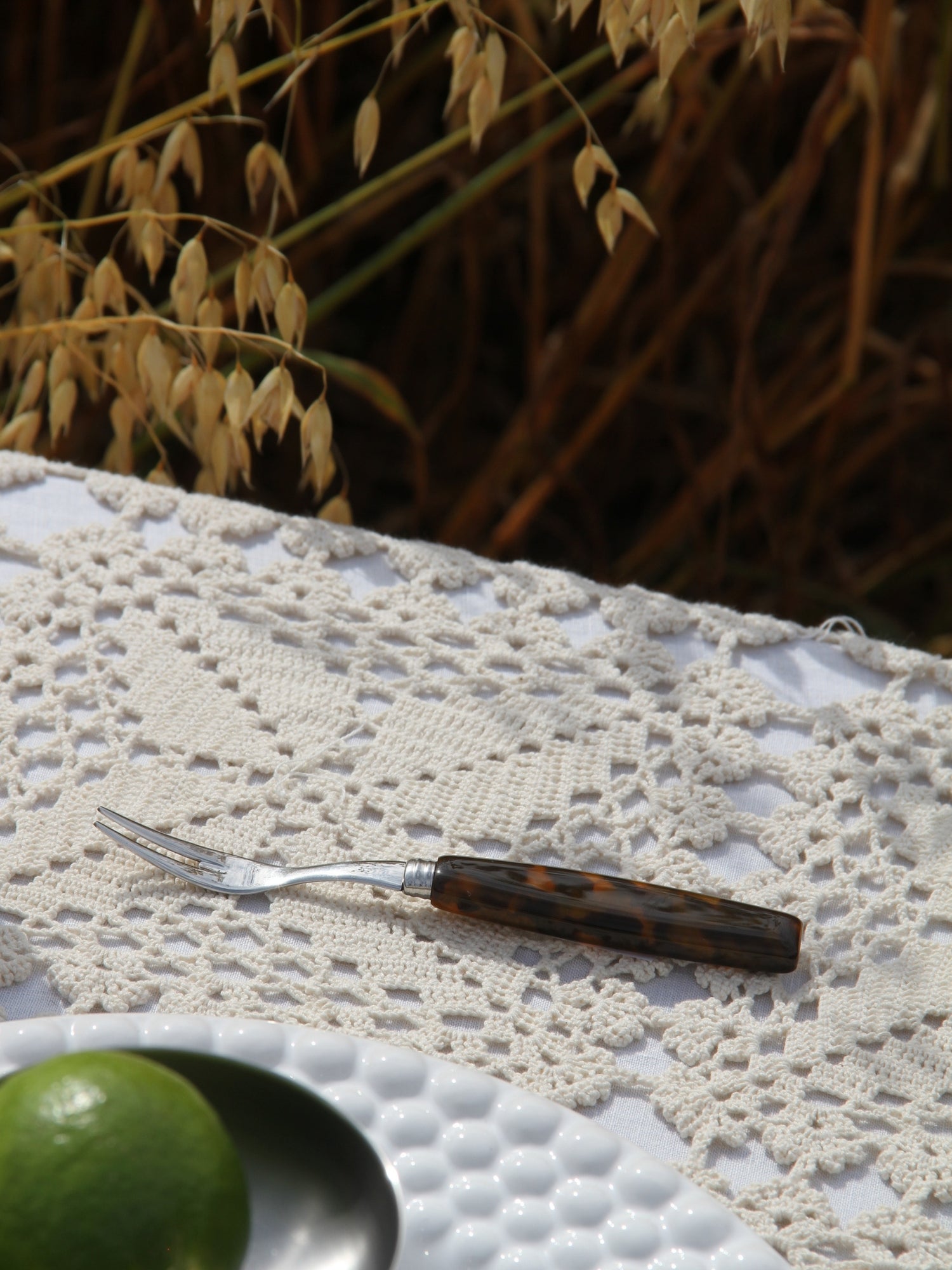 A small serving fork with a tortoiseshell-patterned handle rests on a white lace tablecloth outdoors, with dried grass in the background and part of a green lime visible in the lower left corner.