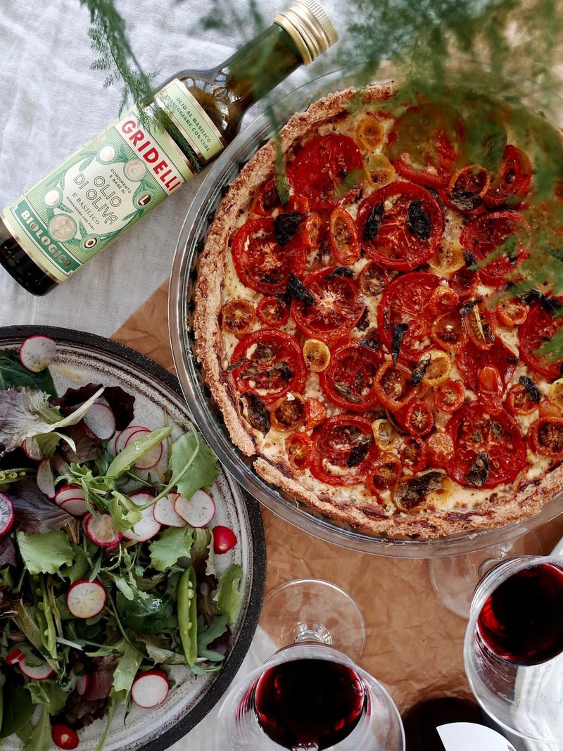 A top-down styled food scene featuring a round tomato and olive oil tart in a glass pie dish, a bowl of mixed salad with radishes, two glasses of red wine, and a bottle of olive oil on a tablecloth.