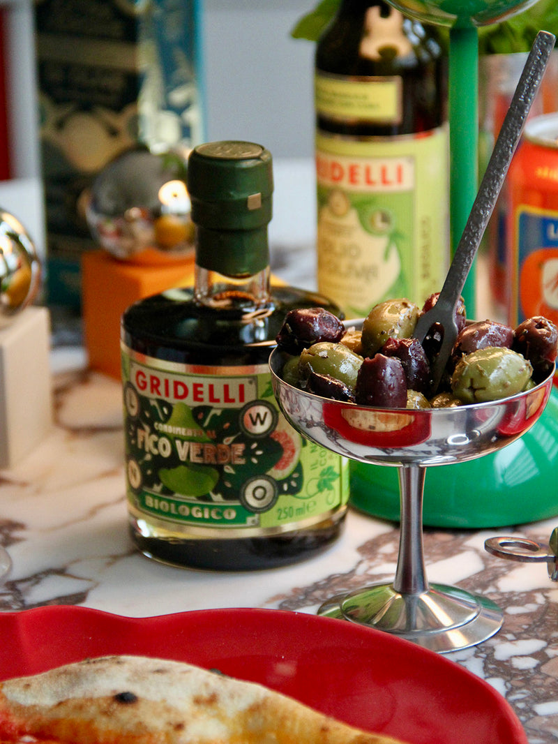 A bottle of Gridelli Pico Verde balsamico stands on a marble countertop beside a stainless steel bowl of stuffed olives with a fork, with other kitchen items blurred in the background.