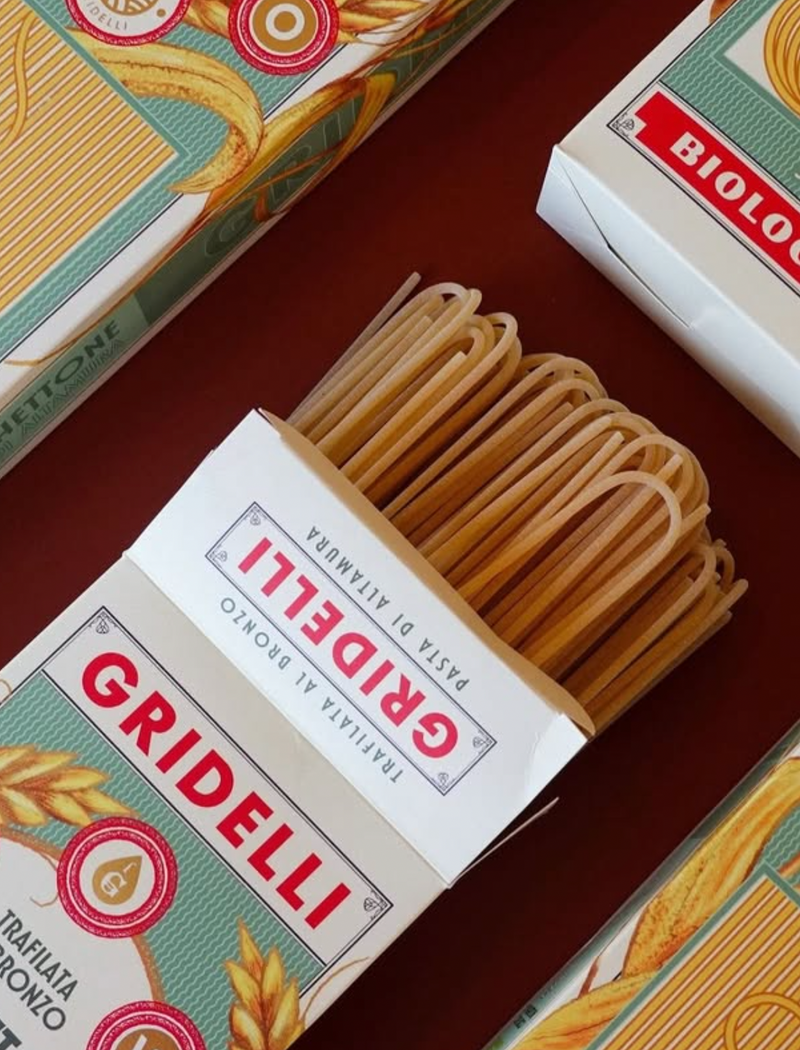 An open box of Gridelli spaghetti pasta on a dark brown surface, with other pasta boxes partially visible around it. The pasta strands are light tan/beige and the packaging is white with red and green branding.