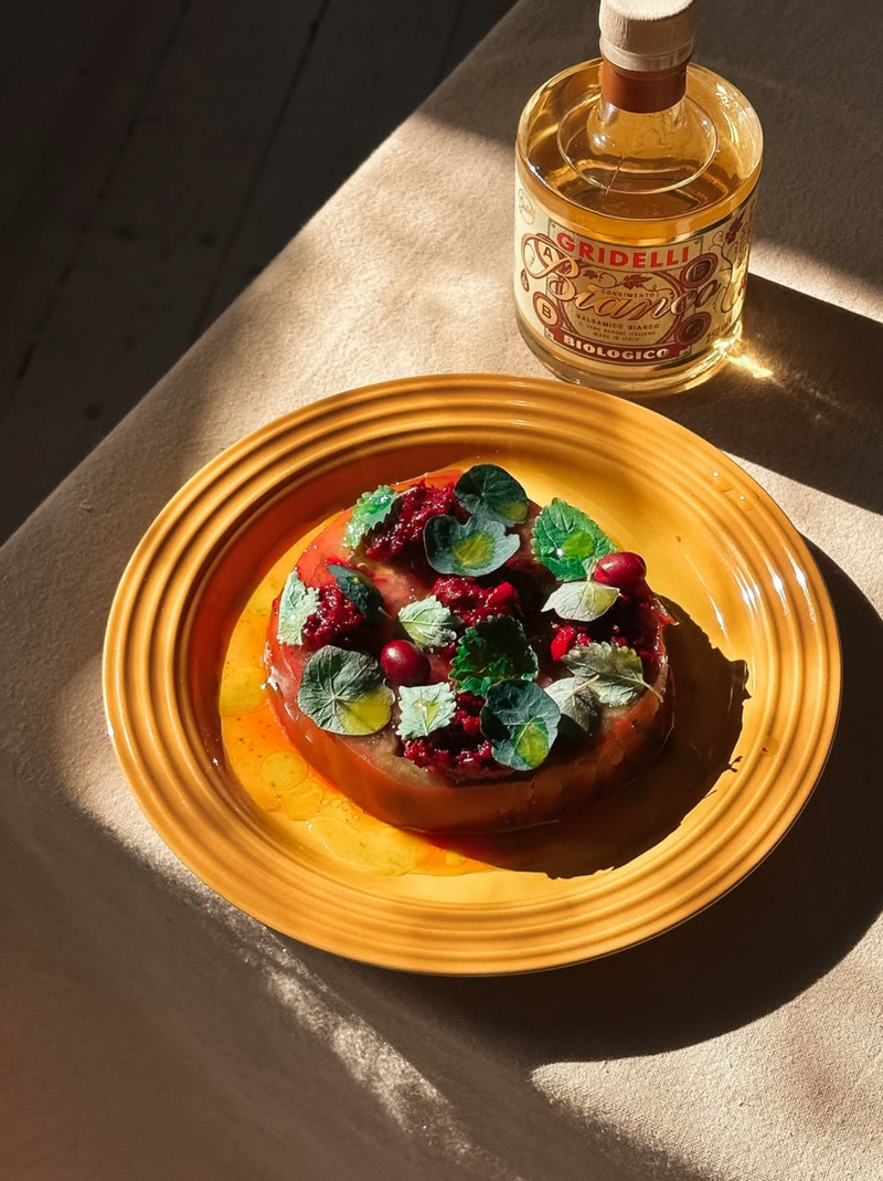 A plated dessert or pastry topped with green leaves and red berries sits on a yellow-orange plate, with a bottle of Brigelli Risotto酒 nearby on a table under strong sunlight.
