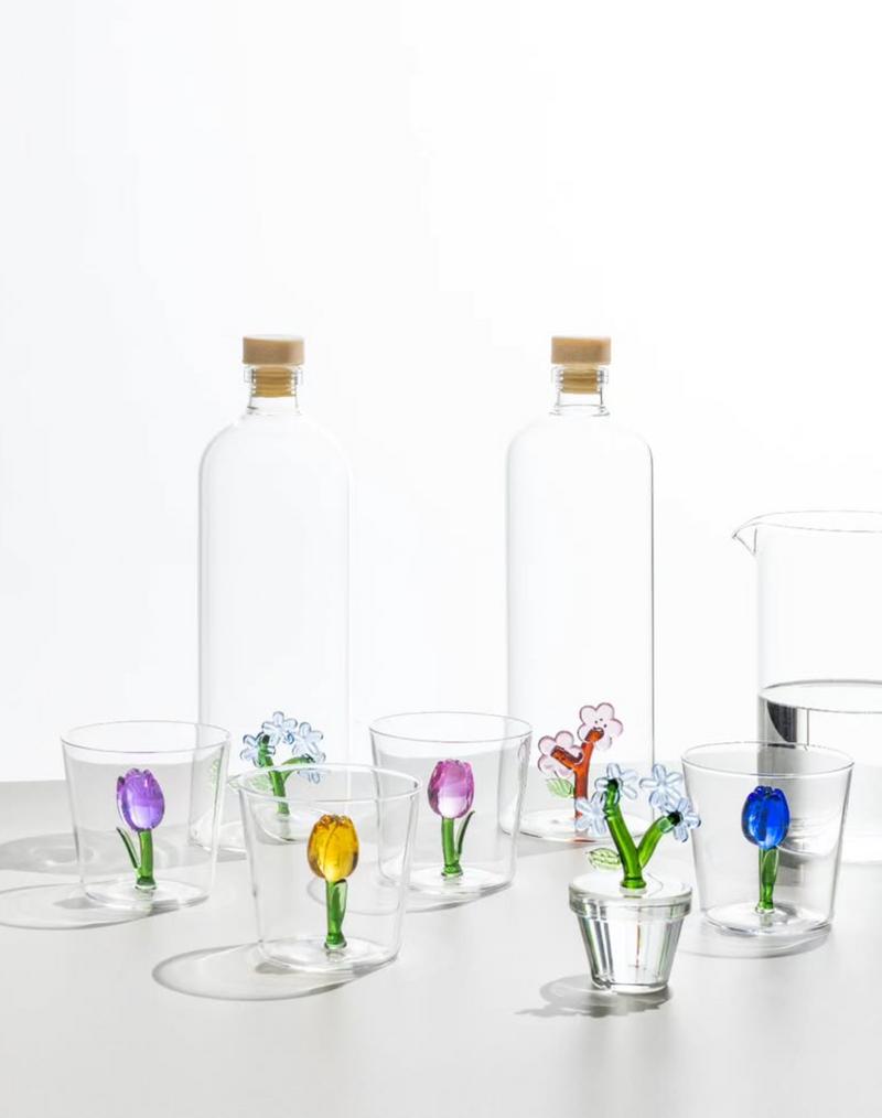 A set of clear glassware including two tall bottles with cork stoppers, several small tumblers, and a pitcher, each decorated with a colorful hand-blown tulip flower in the center, arranged on a white surface against a white background.