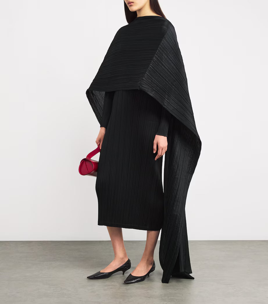 Woman wearing a black dress with a matching cape and holding a red handbag on a plain background