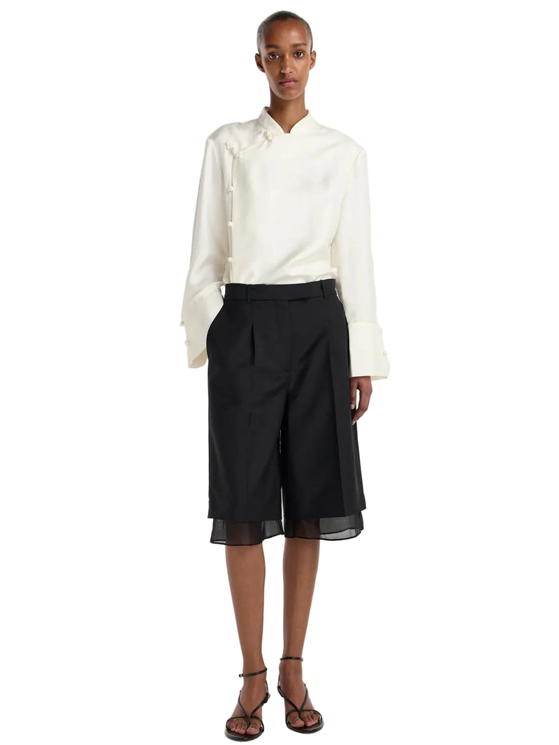 A model stands against a light gray studio background wearing a white long-sleeve shirt with a ruffled collar and front buttons, black high-waisted cropped shorts with a sheer hem, and brown strappy sandals.