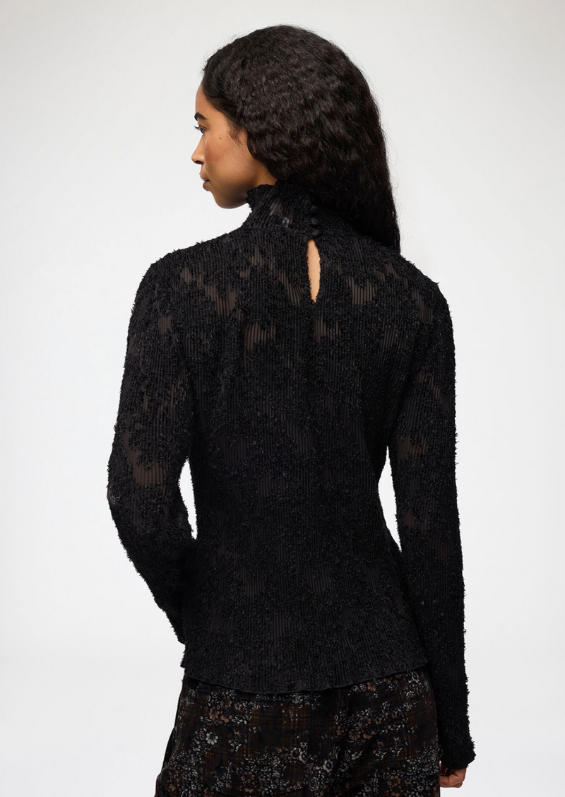 A woman shown from the back wearing a black textured, sheer lace long-sleeve top with a small keyhole cutout at the high neck, set against a plain light gray studio background.