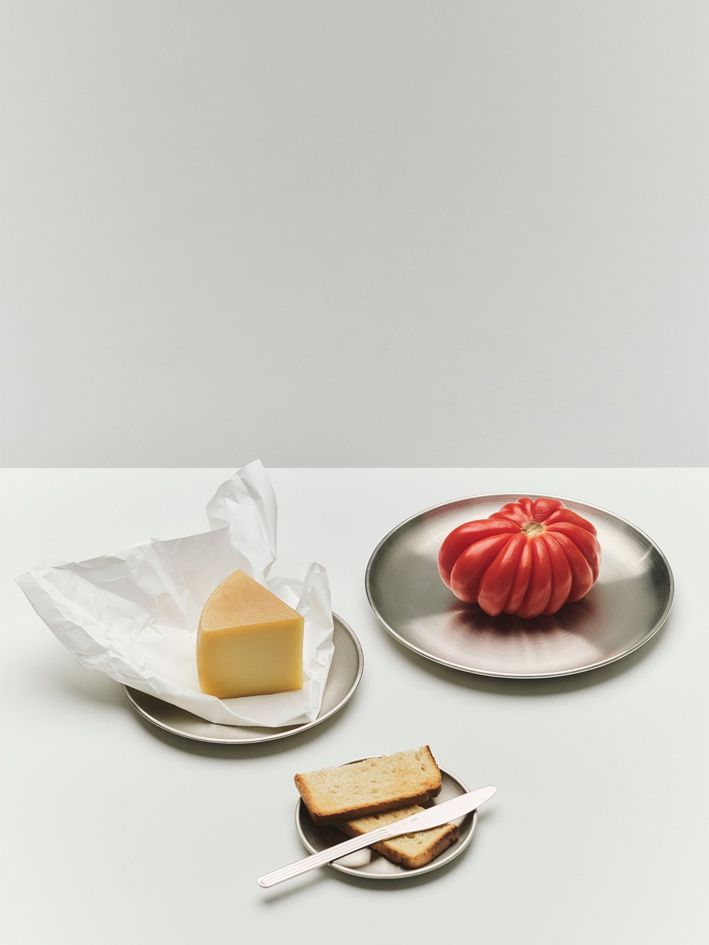 A styled food still life on a light gray surface: a wedge of yellow cheese on white wrapping on shiny metal side plate, a red heirloom tomato on a round shiny metal dinner plate, and two slices of bread with a knife on a small metal plate.
