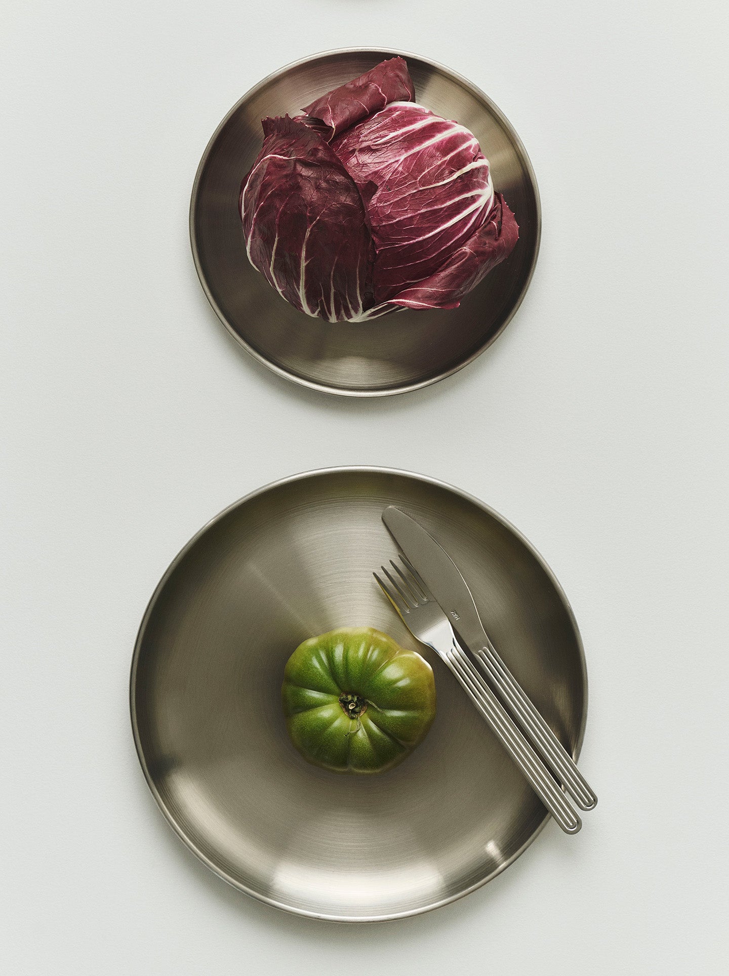 Two round brushed-metal plates on a light gray background. The smaller top plate holds a head of red cabbage, and the larger bottom is a dinner plate holding a green tomato with a fork and knife.