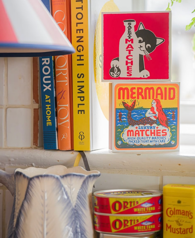 Vintage matchbox and product packaging on a shelf with books and a lamp.