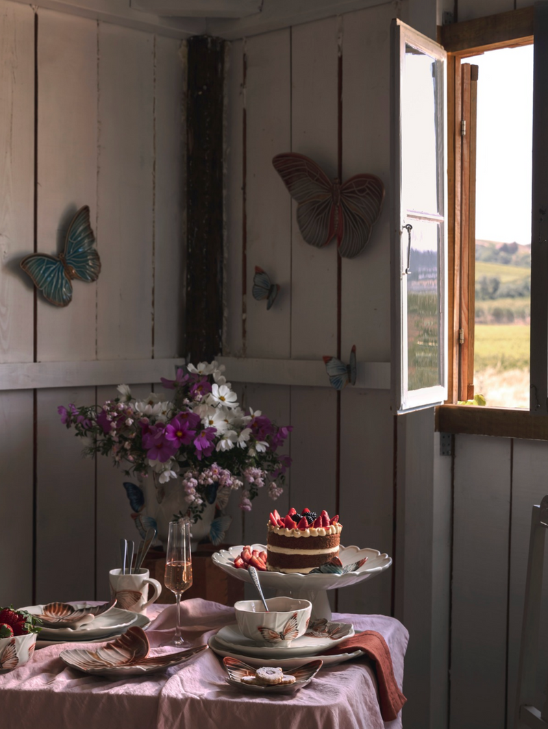 Dining table set with a cake, flowers, and cutlery in a room with wooden walls and a window.